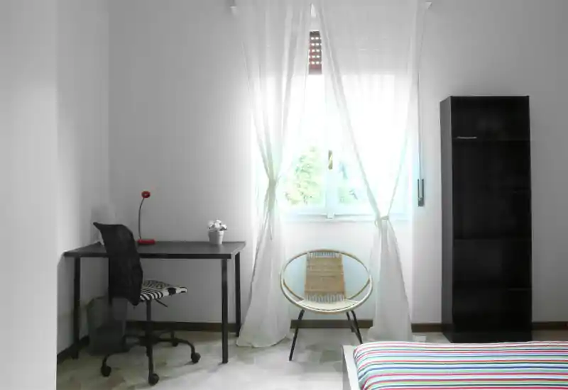 Bright, minimal bedroom with a striped bed in the foreground, a simple desk and chair by a large window letting in abundant natural light.