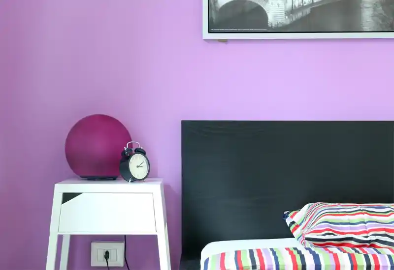 Bright, modern bedroom corner with a black headboard, colorful striped bedding and a white bedside table topped with a round magenta lamp and a classic alarm clock.