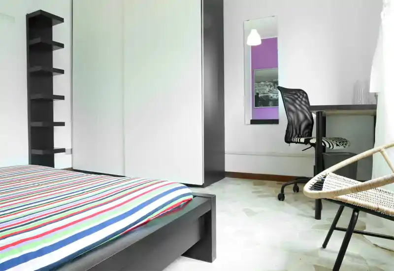 Bright, modern bedroom with a striped double bed in the foreground, built-in wardrobe, desk and chair by the window — clean lines and natural light make it ideal for comfortable stays.