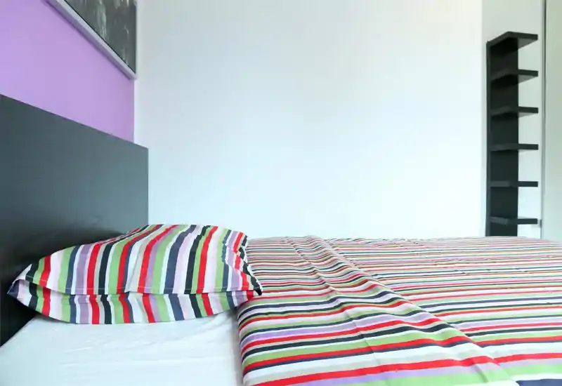 Bright, modern bedroom with a striped duvet and matching pillow on a dark headboard. Minimalist shelving and clean white walls give a fresh, airy feel.
