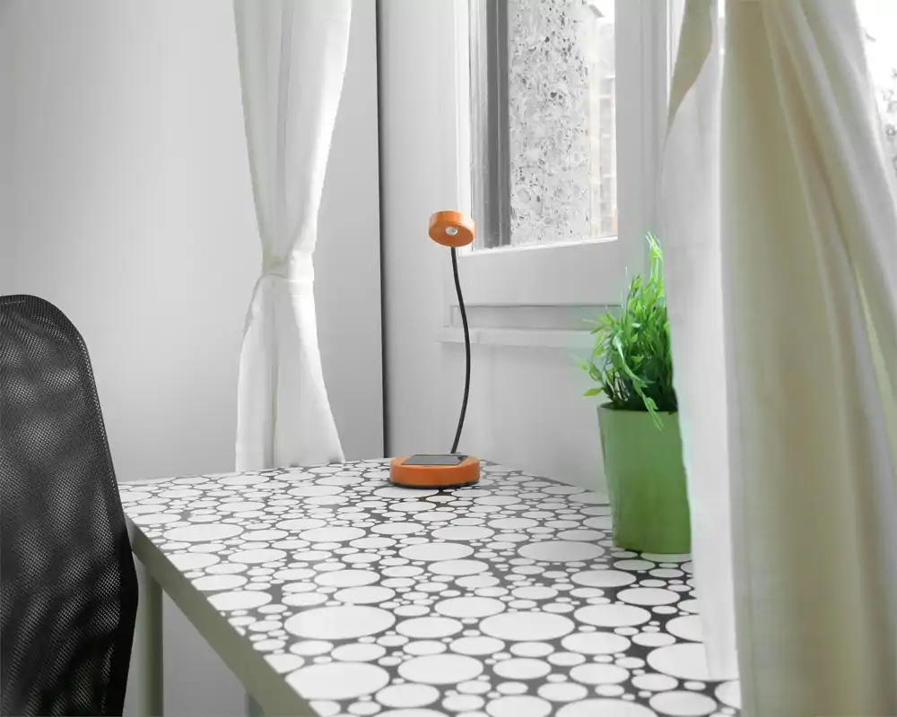 Spazio di lavoro minimalista e luminoso vicino alla finestra con scrivania a pattern, piccola lampada arancione e vaso verde — ideale per lavorare con luce naturale piacevole.