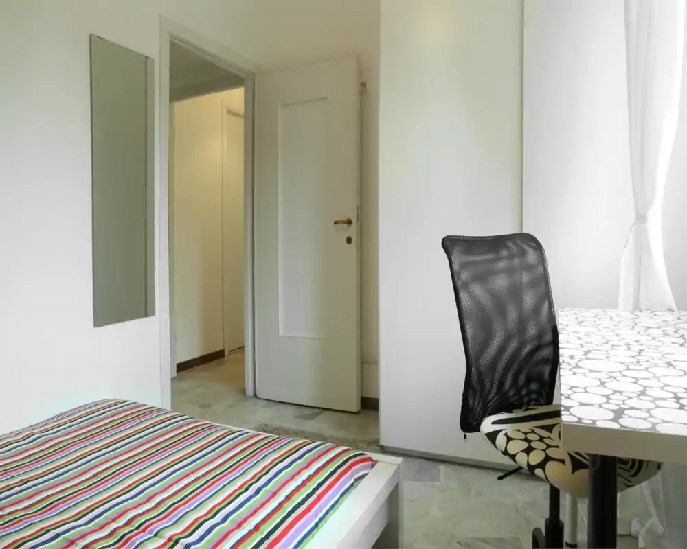 Camera da letto minimalista e luminosa con copriletto a righe, luce naturale da finestra e una scrivania con sedia in rete — semplice, moderna e ben illuminata.