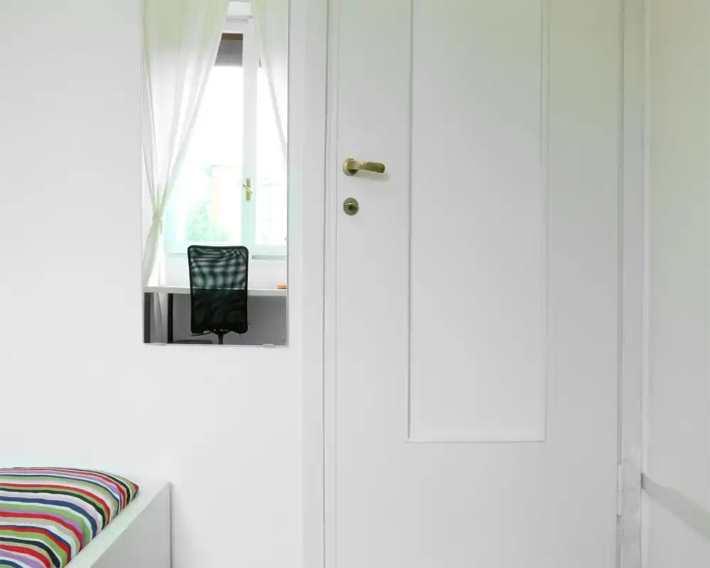Angolo camera da letto luminoso e minimale con un letto a righe e una postazione davanti alla finestra visibile nello specchio; luce naturale nitida e pareti bianche che danno un senso di ariosità.