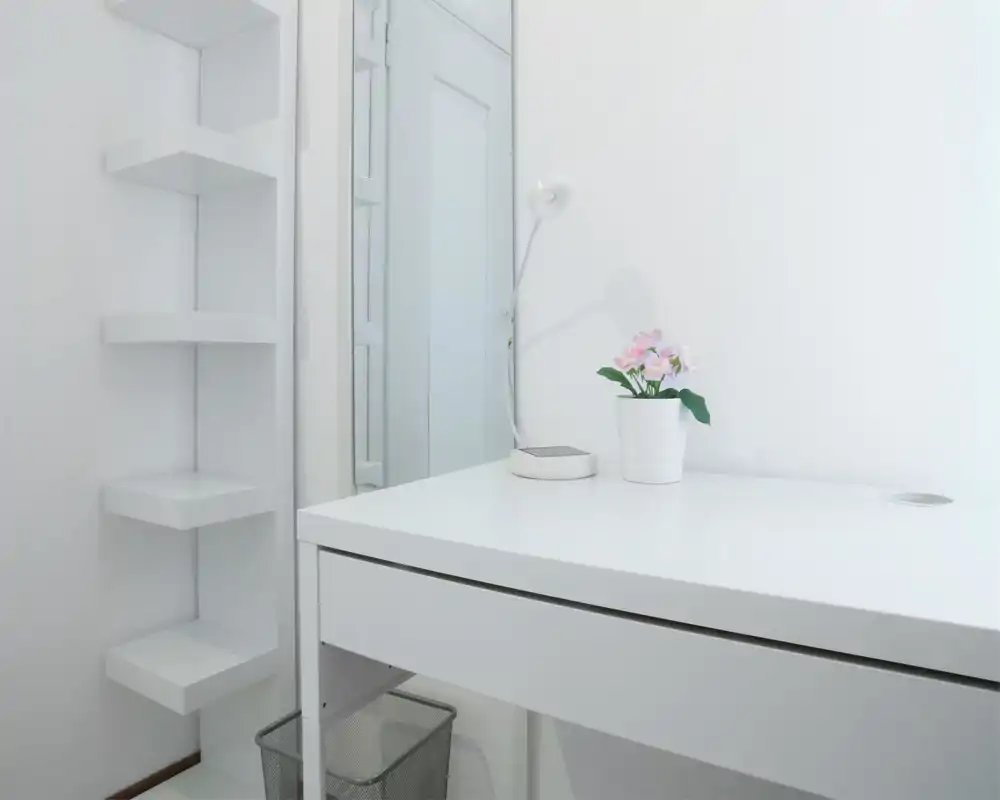 Angolo scrivania minimal bianco e luminoso con lampada e pianta in vaso; ambiente pulito in stile scandinavo ideale come postazione o toeletta.