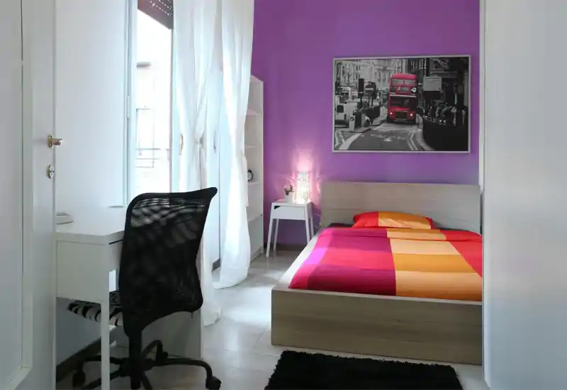Dormitorio moderno y luminoso con cama completa, ropa de cama colorida y un pequeño escritorio junto a la ventana. Luz natural, lámpara de noche y cuadro urbano crean un ambiente fresco y acogedor.