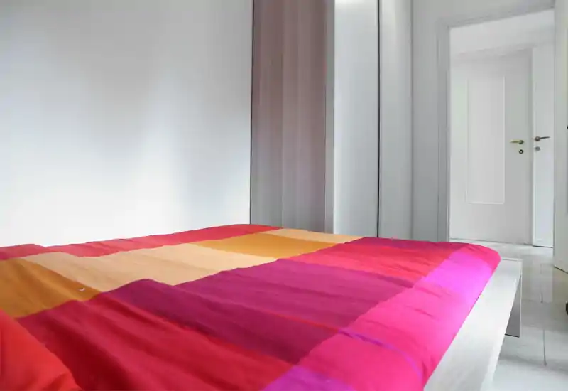 Dormitorio moderno y luminoso con una colcha colorida en primer plano; paredes blancas y armario con espejo que aportan sensación de limpieza y amplitud.