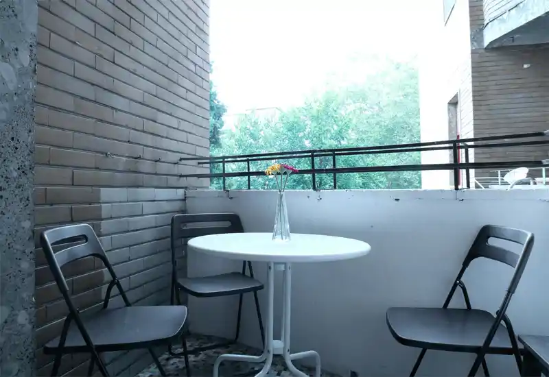 Pequeño balcón privado con mesa redonda blanca, tres sillas plegables y un jarrón con flores; con vistas a los árboles, ideal como zona exterior tranquila.