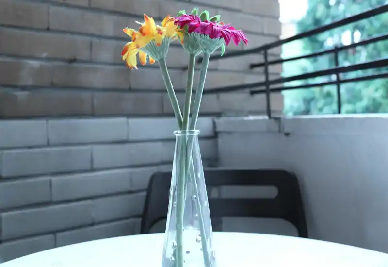 Pequeña escena en balcón con un jarrón de cristal que contiene tres flores artificiales sobre una mesa redonda; pared de ladrillo y barandilla metálica al fondo.