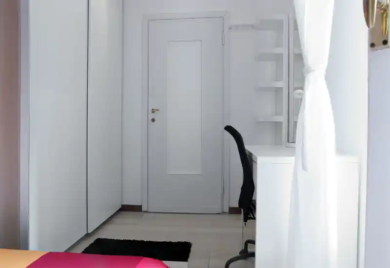 Rincón de dormitorio luminoso con armario blanco, puerta en el centro y un escritorio blanco compacto con silla negra; minimalista y bien iluminado.