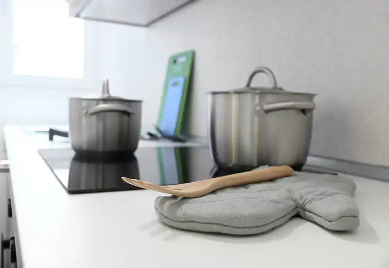 Modern, bright kitchen counter with stainless pots on an induction hob, a wooden spoon and a padded oven mitt in the foreground — a clean, inviting cooking area.