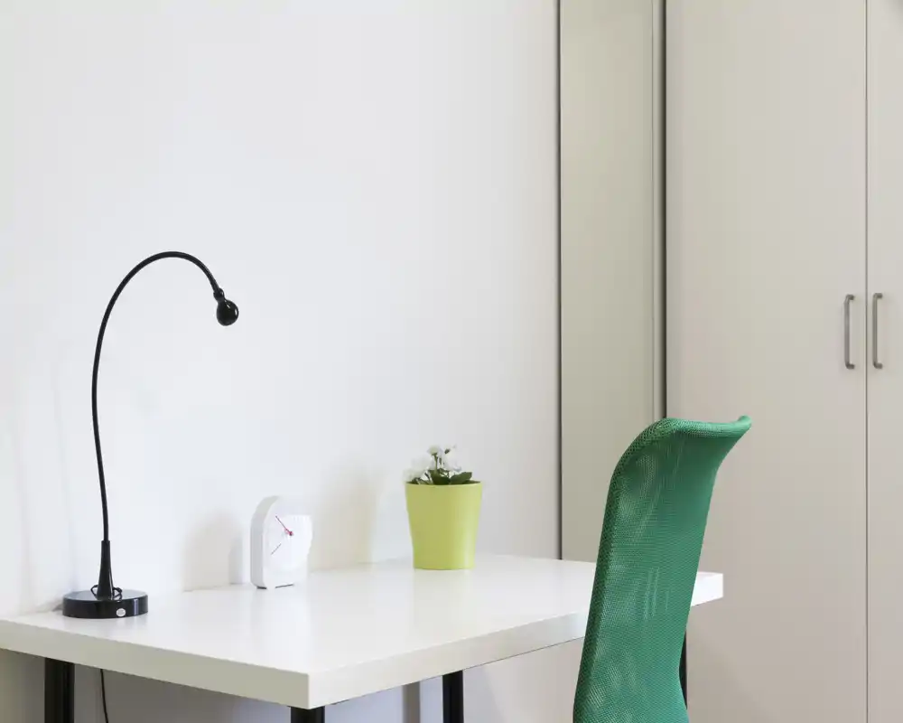 Angolo lavoro minimale e luminoso con scrivania bianca, lampada nera ad arco, piccolo orologio e sedia verde in rete — uno spazio ordinato per studio o smart working.