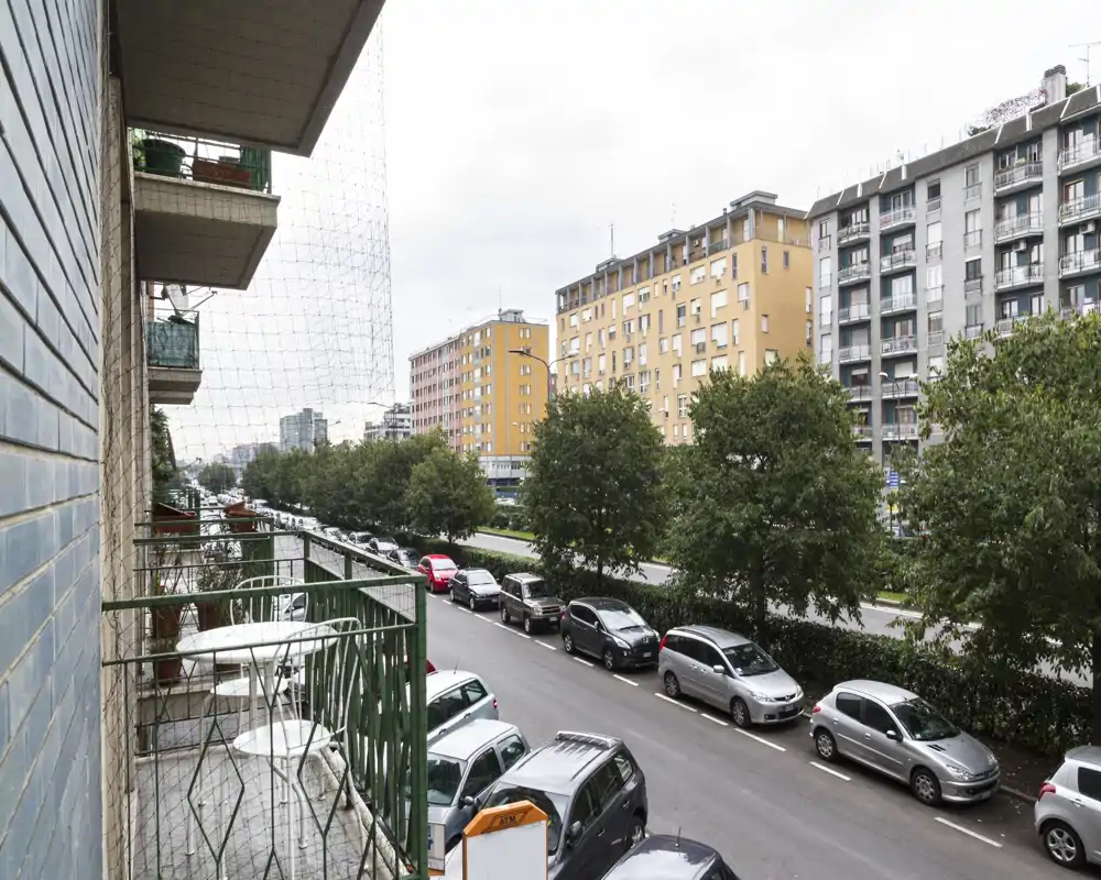 Foto da un balcone verso la strada che mostra un balcone stretto con tavolino e sedie, auto parcheggiate, viale alberato e palazzi residenziali vicini sotto un cielo coperto.