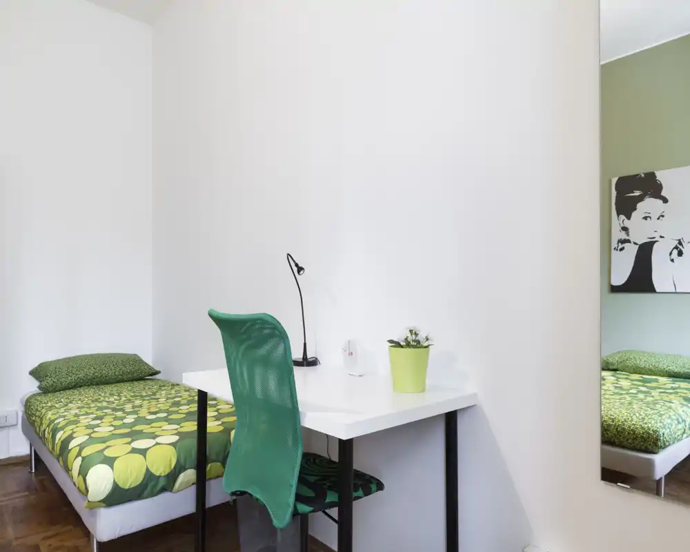Camera da letto singola luminosa con letto con accenti verdi e una scrivania bianca compatta, sedia a rete verde e pianta decorativa — ideale per uno studente o un affitto singolo.