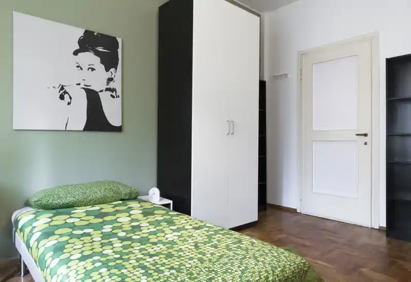 Camera da letto singola luminosa e ordinata con copriletto verde a fantasia, comodino e armadio; quadro minimale e pavimento in legno donano un’atmosfera fresca e accogliente.