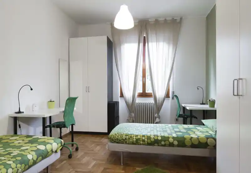 Camera doppia luminosa e ordinata con due letti singoli con biancheria verde a fantasia, due scrivanie con sedie e un armadio — ambiente semplice e adatto a studenti.