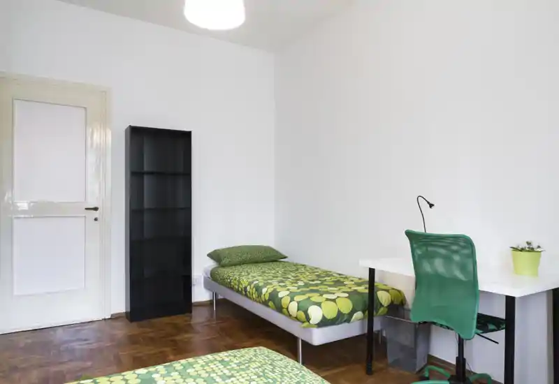 Camera da letto luminosa e minimal con letto singolo con copriletto verde a fantasia, una scrivania semplice con sedia verde e una libreria nera alta — ideale per studenti o affitti brevi.