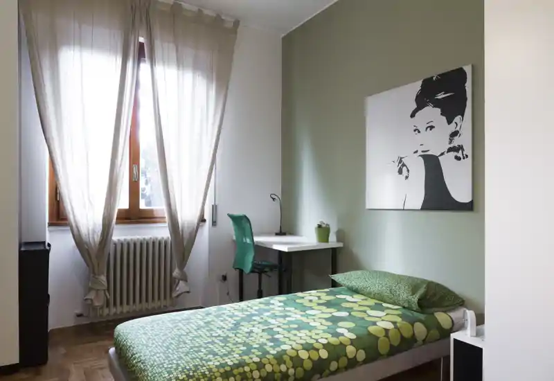 Camera da letto singola luminosa e ordinata con copriletto verde a motivo, una scrivania e sedia vicino alla finestra e una stampa decorativa a parete. La grande finestra con tende leggere dona luce naturale, creando un ambiente accogliente ideale per un annuncio.