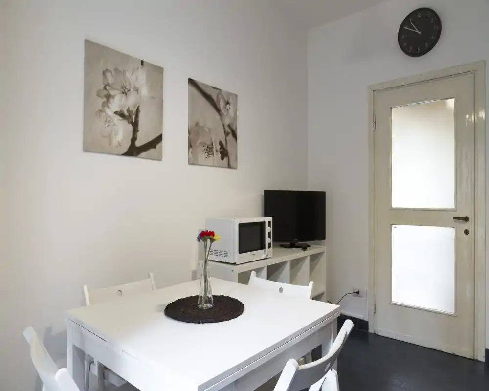 Zona pranzo luminosa e ordinata con tavolo e sedie bianchi, semplici quadri alle pareti e una piccola unità con microonde e TV, perfetta per appartamenti compatti.