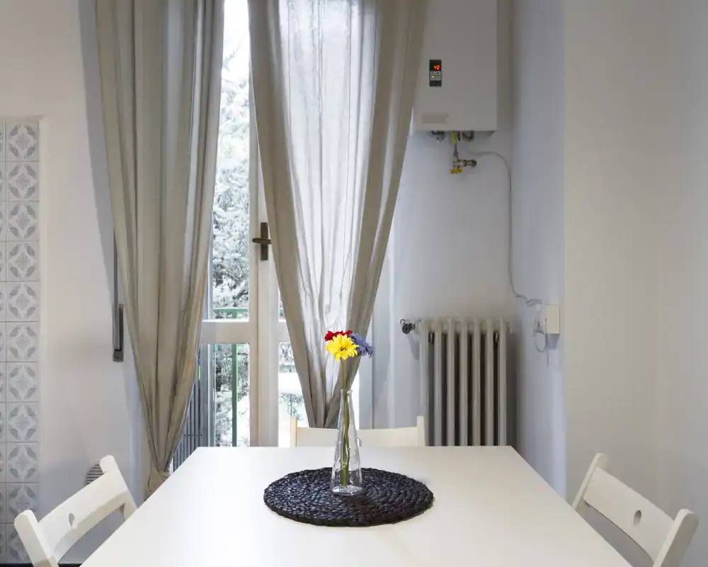 Zona pranzo luminosa e minimale con tavolo bianco, sedie semplici e un piccolo centrotavola floreale davanti a una porta finestra con tende leggere.