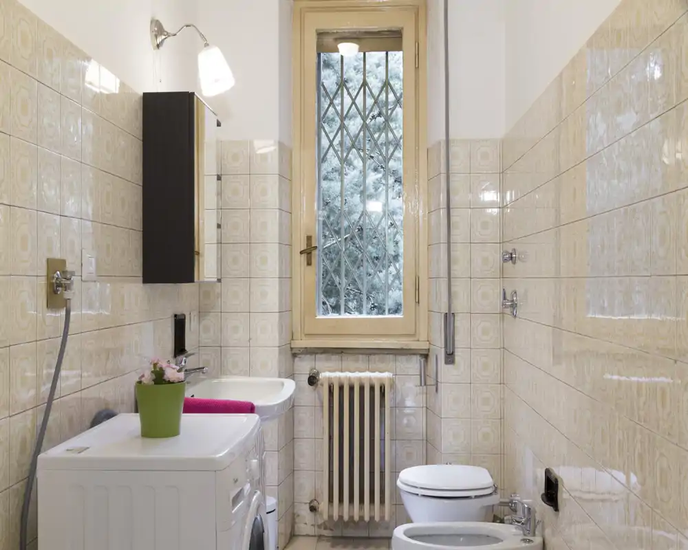 Bagno luminoso e ordinato con lavabo, WC, bidet e lavatrice. La luce dalla finestra alta e le piastrelle decorate donano un aspetto pulito e accogliente.