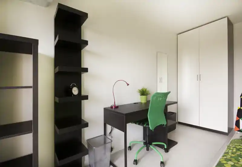 Bright, modern bedroom corner with a small desk and ergonomic green chair next to a wardrobe; minimal decorative touches include a plant and lamp, suitable for a student or single occupant.