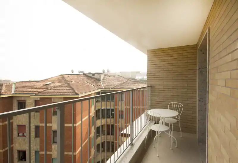 Sunny balcony with a small round table and two metal chairs, overlooking neighboring brick buildings and tiled roofs — a cozy spot for morning coffee.