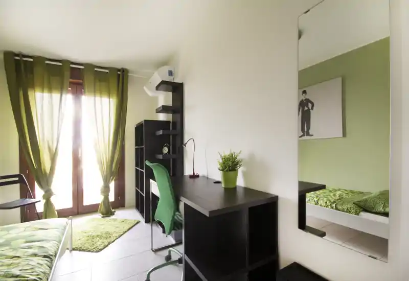 Bright, modern bedroom with green accents: a bed with patterned green bedding, a desk and shelving, and large window doors with green curtains providing abundant natural light.