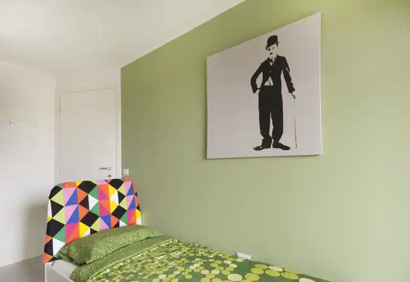 Small, bright bedroom with a single bed featuring a colorful geometric headboard, green patterned bedding and minimalist wall art on a soft green wall.