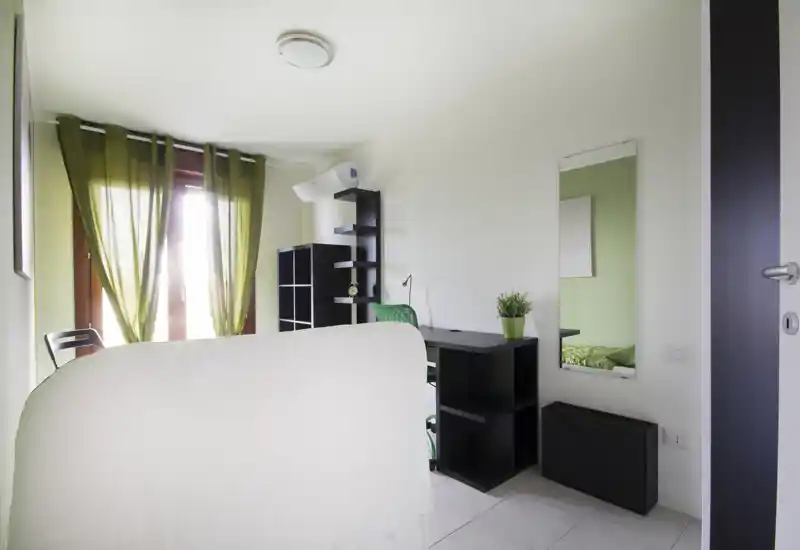 Bright, tidy bedroom corner with a partially visible bed, desk and shelving; green curtains and accents create a calm, modern feel ideal for a single occupant or student.