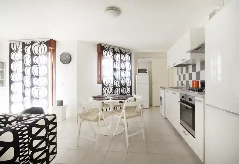 Bright modern open-plan living area with a small dining table and integrated kitchenette; clean white finishes, patterned textiles and abundant natural light create a fresh, inviting space.