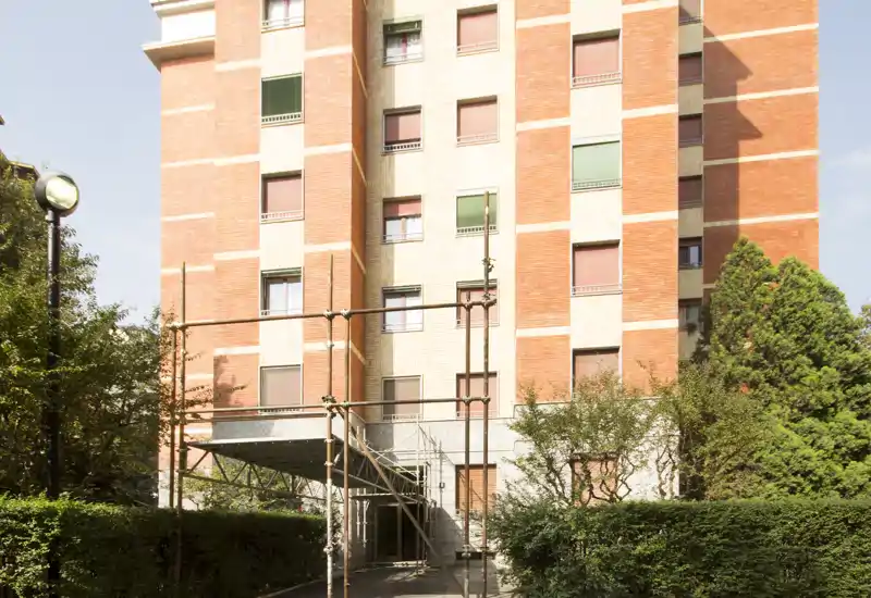 Exterior view of a residential building entrance with scaffolding and landscaped hedges under daylight; shows façade details and surrounding greenery.