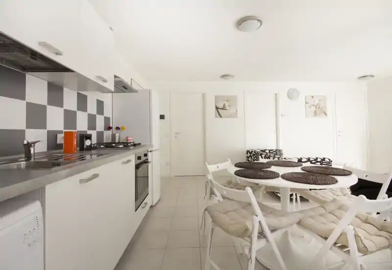 Bright modern kitchen with a long white countertop, built-in oven and a round dining table with cushioned folding chairs—clean, contemporary and inviting for meals.