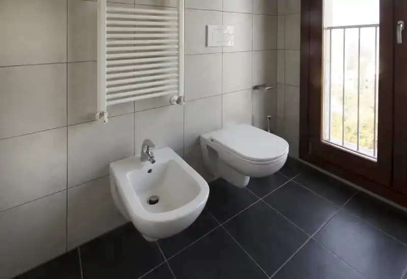 Clean modern bathroom with wall-mounted toilet and bidet, large window providing natural light and tiled walls and floor.