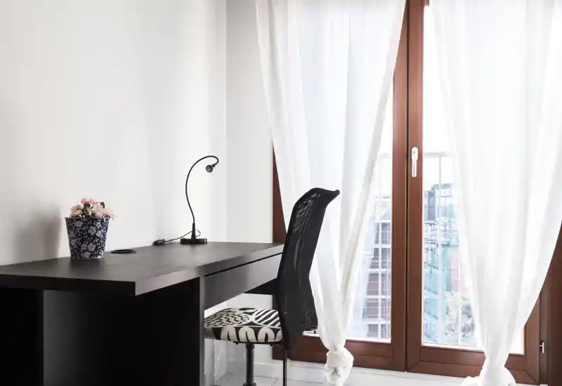 Coin bureau lumineux avec un bureau en bois foncé, chaise à motif, petite lampe et plante en pot à côté de grandes fenêtres voilées blanches laissant entrer la lumière naturelle.