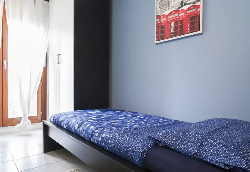 Chambre simple lumineuse avec lit à motifs bleus, cadre de lit sombre et grande fenêtre avec rideaux blancs. Décor épuré et minimaliste, lumière naturelle douce et affiche encadrée au mur.
