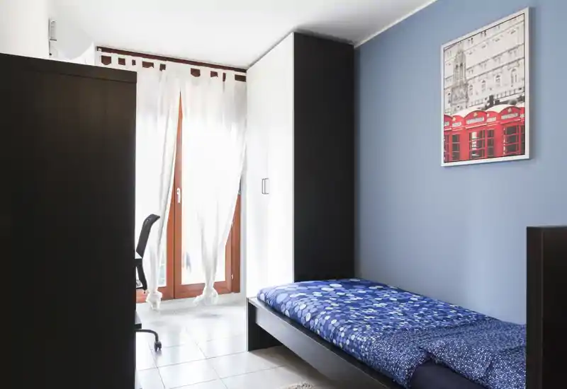 Chambre simple soignée avec lit en bois foncé, linge de lit bleu à motifs et armoire près d'une fenêtre lumineuse ; la lumière naturelle et la décoration minimaliste offrent une atmosphère apaisante.