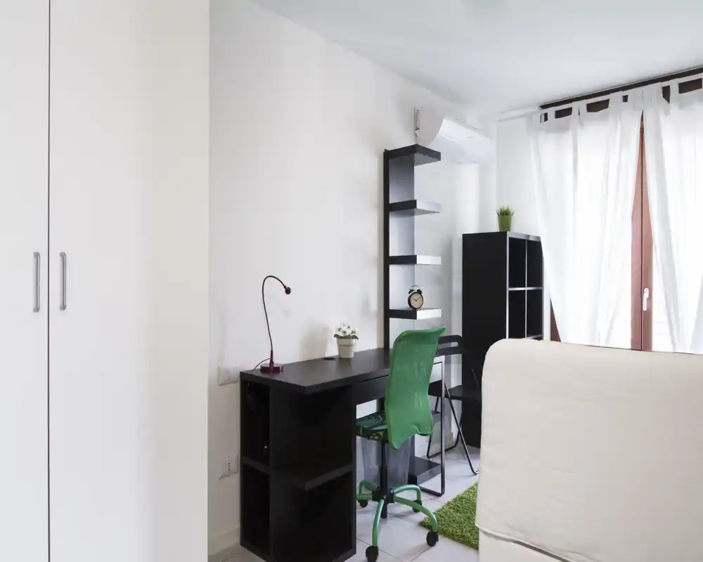 Rincón de dormitorio luminoso y moderno con cama parcialmente visible, escritorio negro con estanterías y silla verde junto a una ventana con cortinas traslúcidas.