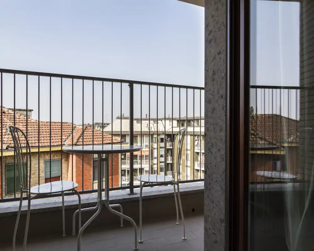 Pequeño balcón amueblado con mesa y dos sillas de metal, tejados de la ciudad visibles más allá de la barandilla y buena luz natural.