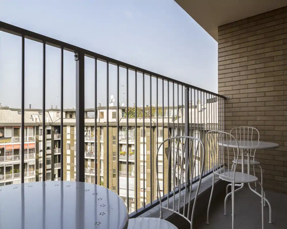 Balcón soleado con barandillas metálicas y una pequeña mesa redonda con sillas, con vistas a los edificios vecinos — ideal para tomar el café por la mañana.