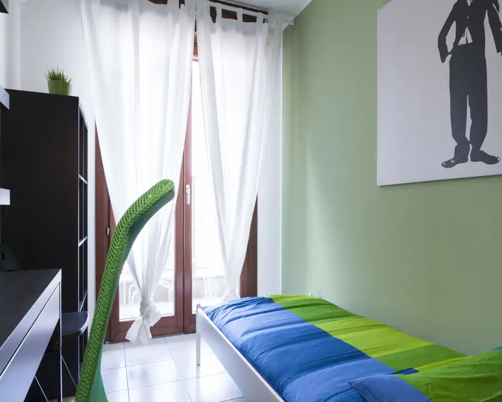 Dormitorio individual luminoso con cama azul y verde, gran ventana con cortinas translúcidas y mobiliario minimalista — ideal para un alquiler compacto y aireado.