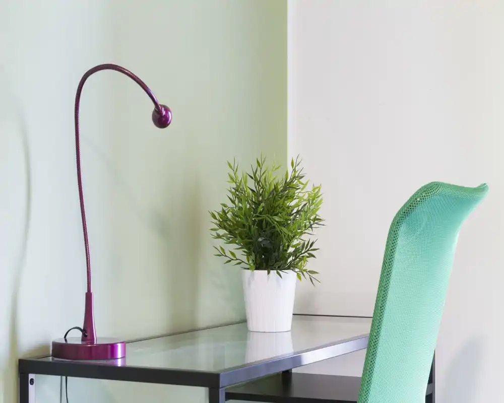 Spazio di lavoro minimale e luminoso con scrivania in vetro, lampada da tavolo fucsia e sedia in rete verde, impreziosito da una pianta in vaso.