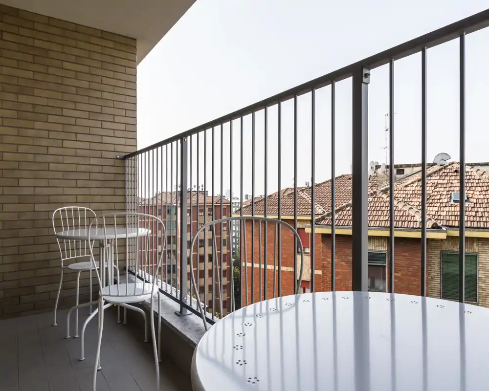 Balcone urbano soleggiato con ringhiera metallica e tavolino e sedie stile bistrot bianche, con vista sugli edifici in mattoni e i tetti in tegole.