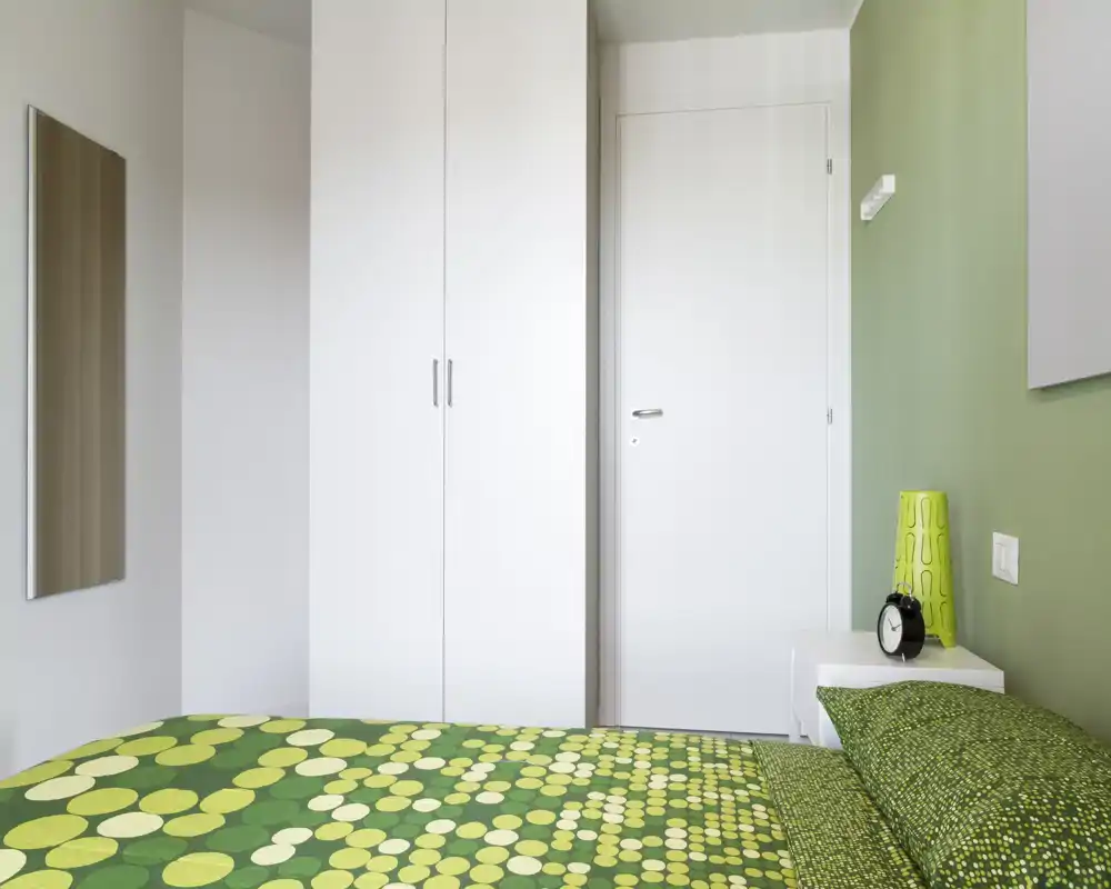 Camera da letto moderna e luminosa con copriletto verde a motivi, armadio bianco e un semplice comodino. La composizione mette in risalto il letto come elemento principale e uno stile pulito e minimal.