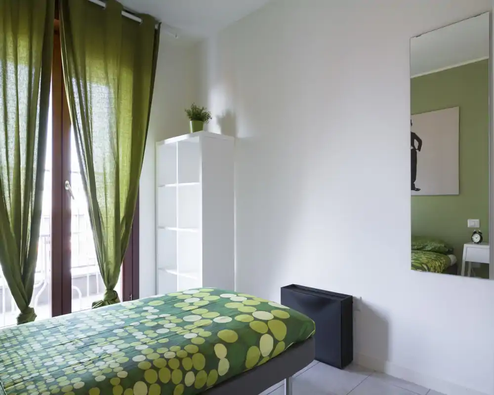 Camera da letto luminosa e ordinata con copriletto verde a fantasia, tende coordinate e uno specchio a tutta altezza che amplifica lo spazio. Mensola semplice e luce naturale dalla porta del balcone rendono l’ambiente arioso e rilassante.