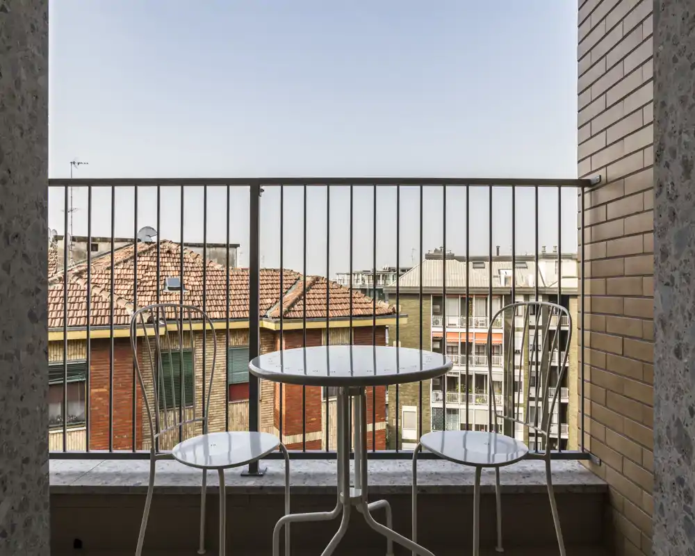 Piccolo balcone soleggiato con tavolino rotondo e due sedie in metallo, con vista sui tetti dei palazzi vicini — perfetto per il caffè mattutino.