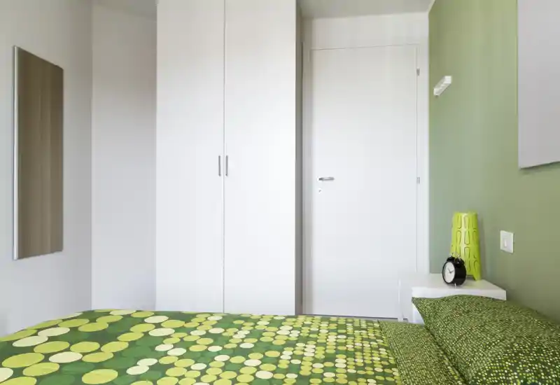 Camera da letto moderna e luminosa con copriletto verde a motivi, armadio bianco e un semplice comodino. La composizione mette in risalto il letto come elemento principale e uno stile pulito e minimal.