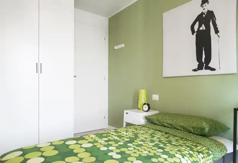 Camera da letto luminosa e moderna con parete verde e biancheria a fantasia verde; comodino con lampada e sveglia e un grande armadio per riporre.