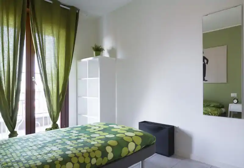 Camera da letto luminosa e ordinata con copriletto verde a fantasia, tende coordinate e uno specchio a tutta altezza che amplifica lo spazio. Mensola semplice e luce naturale dalla porta del balcone rendono l’ambiente arioso e rilassante.
