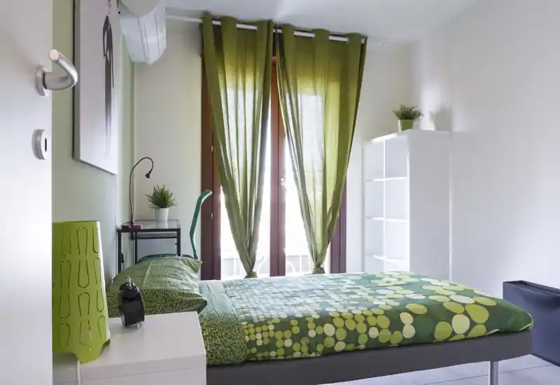 Camera da letto moderna e luminosa con accenti verdi. Il letto matrimoniale ha una biancheria verde a fantasia, un comodino con lampada e ampie finestre con tende verdi che diffondono luce naturale.