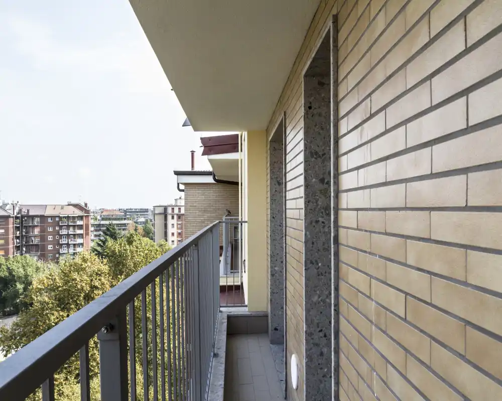 Sunny narrow balcony with metal railing, brick wall and view over nearby residential buildings and trees — ideal for morning coffee or light outdoor seating.